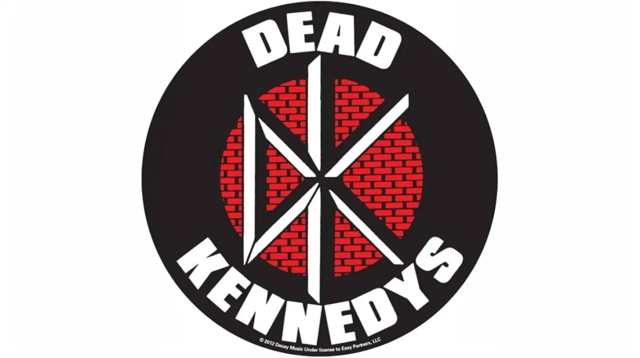 黒い円形の背景に、白文字で「DEAD KENNEDYS」と書かれたロゴ。中央には赤いレンガ模様の円があり、その上に白く鋭角的なDKの文字がデザインされている。