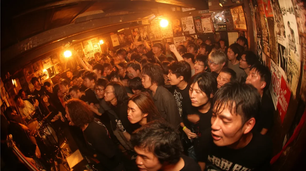 ポスターが所狭しと貼られた狭く薄暗い日本のライブハウスで、熱狂的な観客が密集してバンドの演奏に盛り上がっている様子を魚眼レンズで撮影した写真。ドラマーが左奥に見え、観客は拳を上げたり、叫んだりしている。