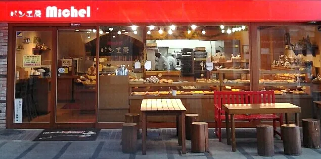 石窯パン工房ミッシェルの盛岡肴町店の外観。赤い看板とガラス張りの店内からパンが並ぶ様子が見え、店先にはテーブル席が設置されている。