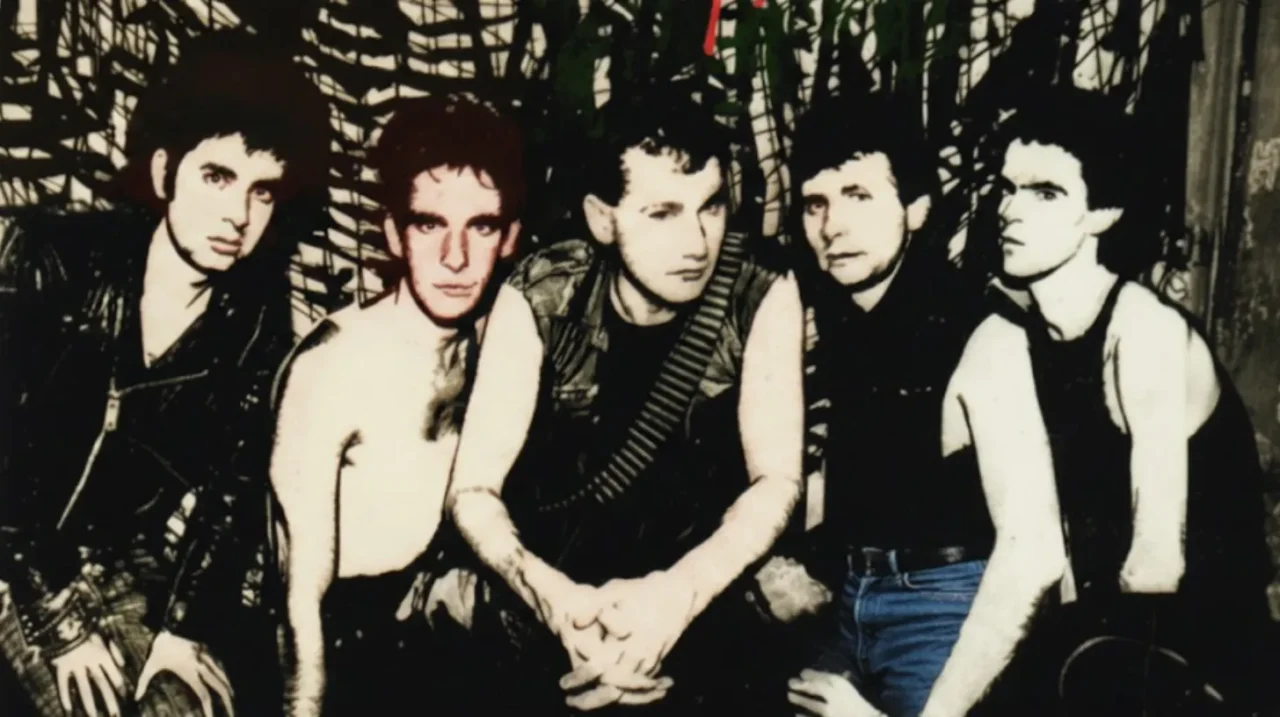 暗めの背景の前で5人の男性が横並びで座っているように配置されている。白黒に近い加工が施され、赤みのある色調が部分的に加えられたアート風の写真。革ジャンやノースリーブなどパンクスタイルの衣服を着た人物が多く、全体的に粗さと緊張感を感じさせるビジュアル。