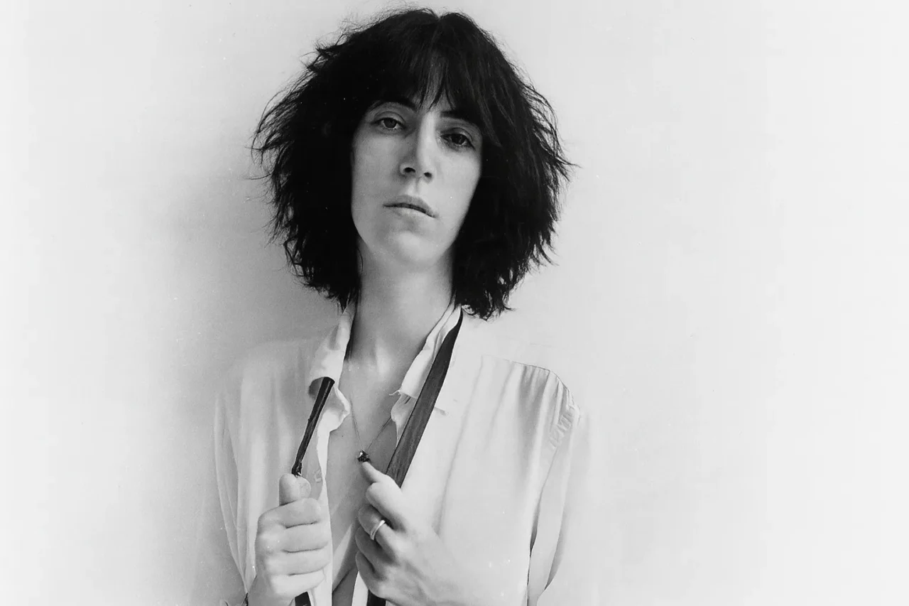 白いシャツを着て黒いネクタイのようなものを肩にかけた、若き日のパティ・スミスの白黒写真。彼女はカメラをまっすぐに見つめている。