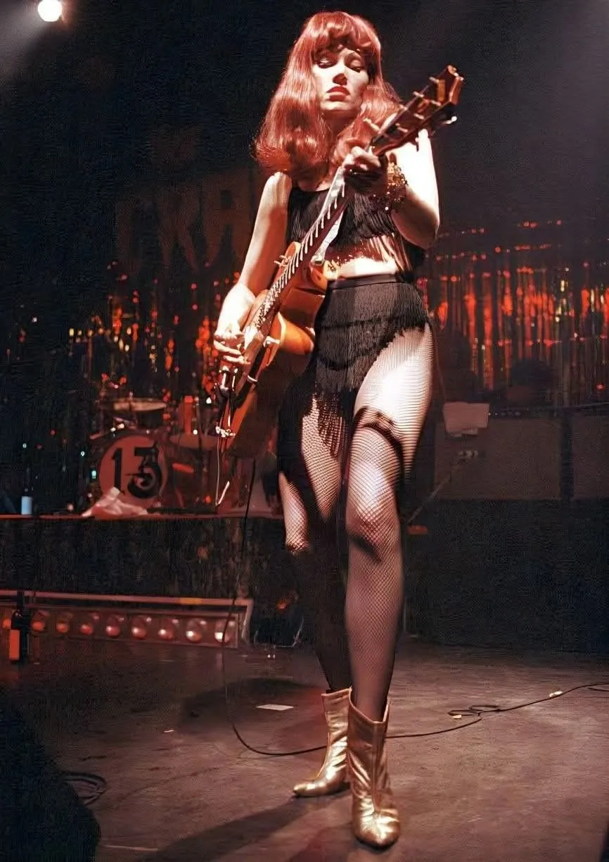 ステージ上でグレッチのホロウボディ・ギターを演奏するザ・クランプスのギタリスト、ポイズン・アイビー。赤い髪、黒いフリンジの衣装、網タイツ、ゴールドのブーツという出で立ち。
